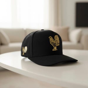 May include: A black baseball cap featuring a gold rooster emblem on the front and side. The cap has a curved brim and a structured design. The rooster is a detailed, stylized illustration.