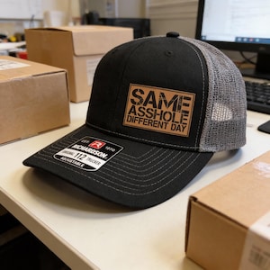 May include: Black trucker hat with a gray mesh back and a curved brim. The hat features a brown square patch with the text "SAME ASSHOLE DIFFERENT DAY". The Richardson 112 hat is adjustable.