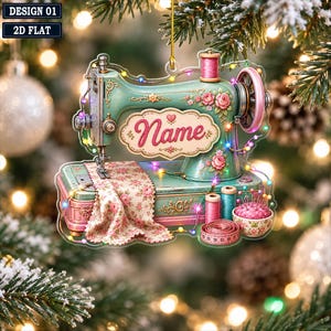 Op de afbeelding: Een plat, hangend ornament in de vorm van een vintage naaimachine in groenblauw en roze met bloemmotieven. Het bevat een centraal plaatje met de tekst 'Name', een garenklosje, een speldenkussen en een meetlint, alles omwikkeld met lichtjes.