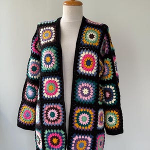 Op de afbeelding: Een handgemaakt, opengebreid vest. De zwarte jas heeft kleurrijke granny squares in tinten roze, geel, blauw en groen. Het vest wordt gepresenteerd op een mannequin tegen een effen witte achtergrond.