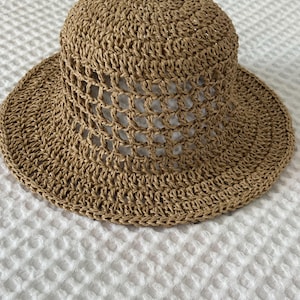 May include: A tan-colored sun hat crafted from woven straw material. The hat features a rounded crown with a wide, flat brim. The middle section of the crown displays an open, grid-like crochet pattern, while the top and brim are tightly woven.