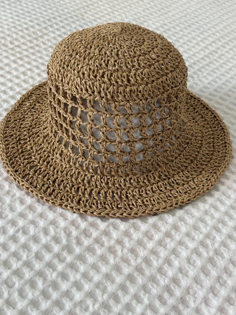 May include: A tan-colored sun hat crafted from woven straw material. The hat features a rounded crown with a wide, flat brim. The middle section of the crown displays an open, grid-like crochet pattern, while the top and brim are tightly woven.