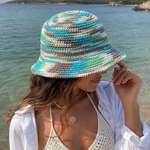 Può includere: Un cappello a secchiello all'uncinetto con un motivo variegato di filato azzurro, turchese brillante, verde salvia e marrone tenue. Il cappello presenta una corona arrotondata e una tesa spiovente, realizzato con un punto denso e strutturato.