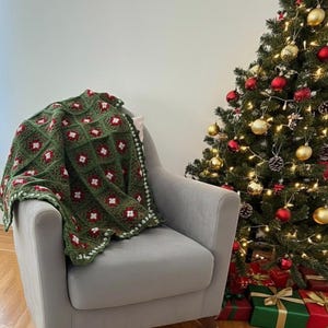 May include: A green and red crocheted blanket draped over a gray armchair. The blanket features a repeating pattern of red squares on a green background. A Christmas tree with ornaments and wrapped gifts is in the background.