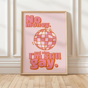 May include: A framed pink print with the text "No money. and also I'm still gay." The print features a disco ball graphic in shades of pink and orange. The frame is a light wood colour.