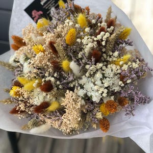 Peut inclure: Un bouquet de fleurs séchées aux tons jaunes, bruns, blancs et violets, enveloppé dans du papier blanc. L'arrangement comprend diverses textures et formes, avec une petite étiquette noire attachée.