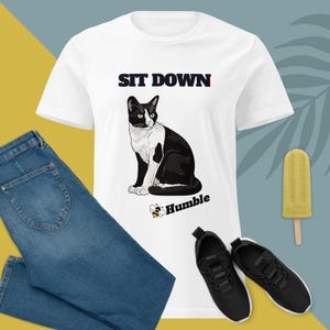 Op de afbeelding: Wit T-shirt met een zwart-witte kattenillustratie en de tekst "SIT DOWN" boven de kat en "Humble" eronder. Het shirt wordt gecombineerd met een blauwe jeans en zwarte sneakers. Een gele ijslolly is ook zichtbaar in de afbeelding.