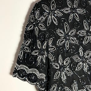 May include: A black top with short sleeves, embellished with white floral beadwork. The design showcases detailed patterns of beaded flowers and leaves, offering a textured and elegant appearance. The sleeves are finished with a scalloped trim.