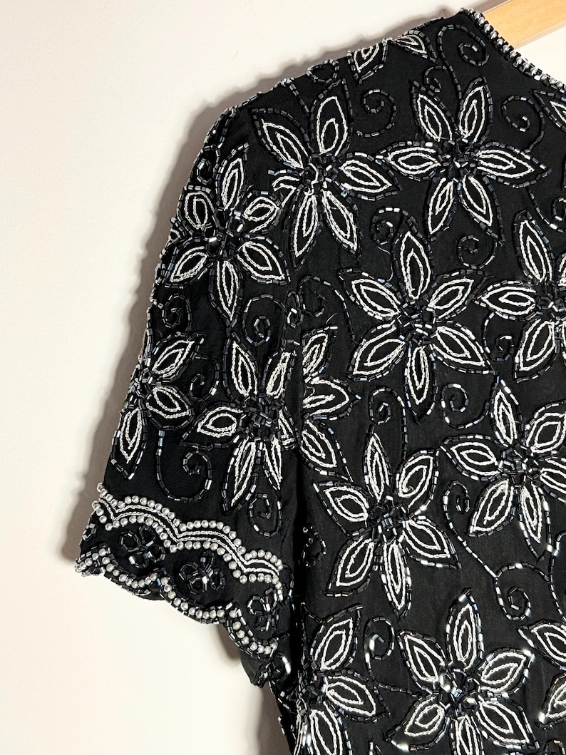 May include: A black top with short sleeves, embellished with white floral beadwork. The design showcases detailed patterns of beaded flowers and leaves, offering a textured and elegant appearance. The sleeves are finished with a scalloped trim.