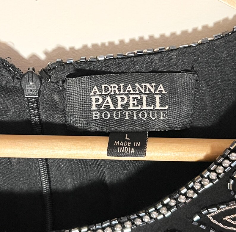 May include: Close-up of a black Adrianna Papell Boutique dress. The label reads "Adrianna Papell Boutique" and "Made in India". The dress has a zipper and is embellished with beads. The dress is size L.