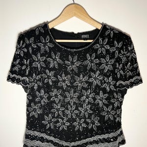May include: Black beaded top with a floral design. The short-sleeved top features a round neckline and a scalloped hem. The floral pattern is made of white beads and sequins. The top is hanging on a wooden hanger.