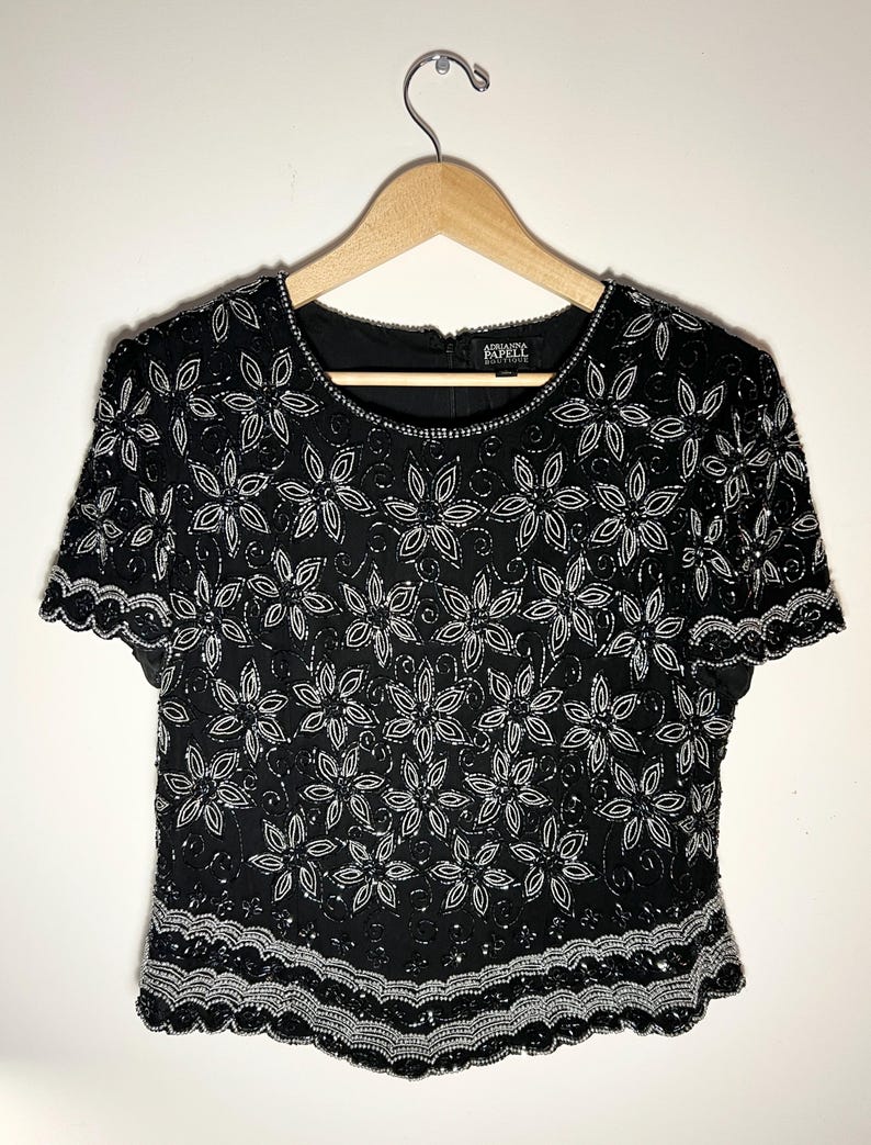 May include: Black beaded top with a floral design. The short-sleeved top features a round neckline and a scalloped hem. The floral pattern is made of white beads and sequins. The top is hanging on a wooden hanger.