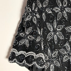 May include: Black short-sleeved top with detailed floral beadwork. The design showcases large, stylised flowers outlined in silver beads, with smaller silver beads filling the petals. The hem and sleeve edges have a scalloped trim.