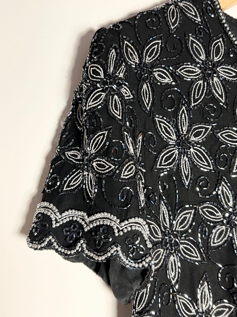May include: Black short-sleeved top with detailed floral beadwork. The design showcases large, stylised flowers outlined in silver beads, with smaller silver beads filling the petals. The hem and sleeve edges have a scalloped trim.
