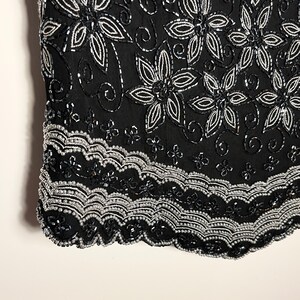 May include: Black top adorned with silver and black beadwork. The design includes floral patterns and a scalloped hem. The intricate beadwork creates a textured effect. The top's design is detailed and elegant, suitable for evening wear.