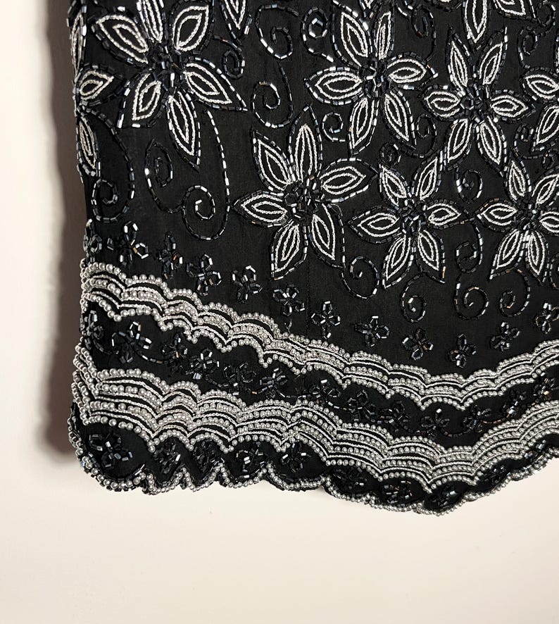 May include: Black top adorned with silver and black beadwork. The design includes floral patterns and a scalloped hem. The intricate beadwork creates a textured effect. The top's design is detailed and elegant, suitable for evening wear.