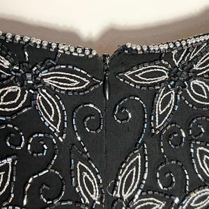 May include: Close-up of a black garment with detailed beadwork. The design includes white and silver beaded floral and leaf patterns, and a zipper closure. The neckline is trimmed with small, silver beads.