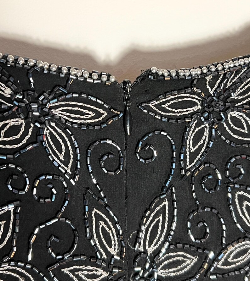 May include: Close-up of a black garment with detailed beadwork. The design includes white and silver beaded floral and leaf patterns, and a zipper closure. The neckline is trimmed with small, silver beads.