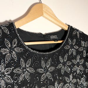 May include: Black dress with floral beadwork. The dress has a round neckline and short sleeves. The floral design is made of silver beads and sequins. The dress is hanging on a wooden hanger. The label reads "Adrianna Papell Boutique."