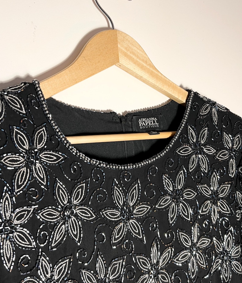 May include: Black dress with floral beadwork. The dress has a round neckline and short sleeves. The floral design is made of silver beads and sequins. The dress is hanging on a wooden hanger. The label reads "Adrianna Papell Boutique."