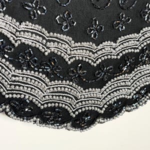 May include: Close-up of black fabric with detailed beadwork. The design incorporates silver and black beads in floral and swirling patterns. The edges are scalloped with a row of silver beads.
