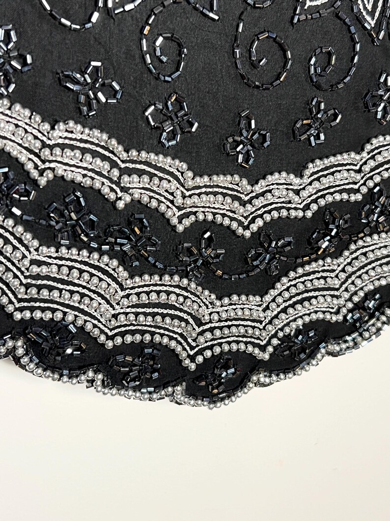 May include: Close-up of black fabric with detailed beadwork. The design incorporates silver and black beads in floral and swirling patterns. The edges are scalloped with a row of silver beads.