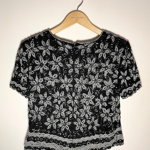 May include: Black beaded top with white floral design. The short-sleeved top is embellished with sequins and beads, featuring a scalloped hemline. The top is hanging on a wooden hanger.