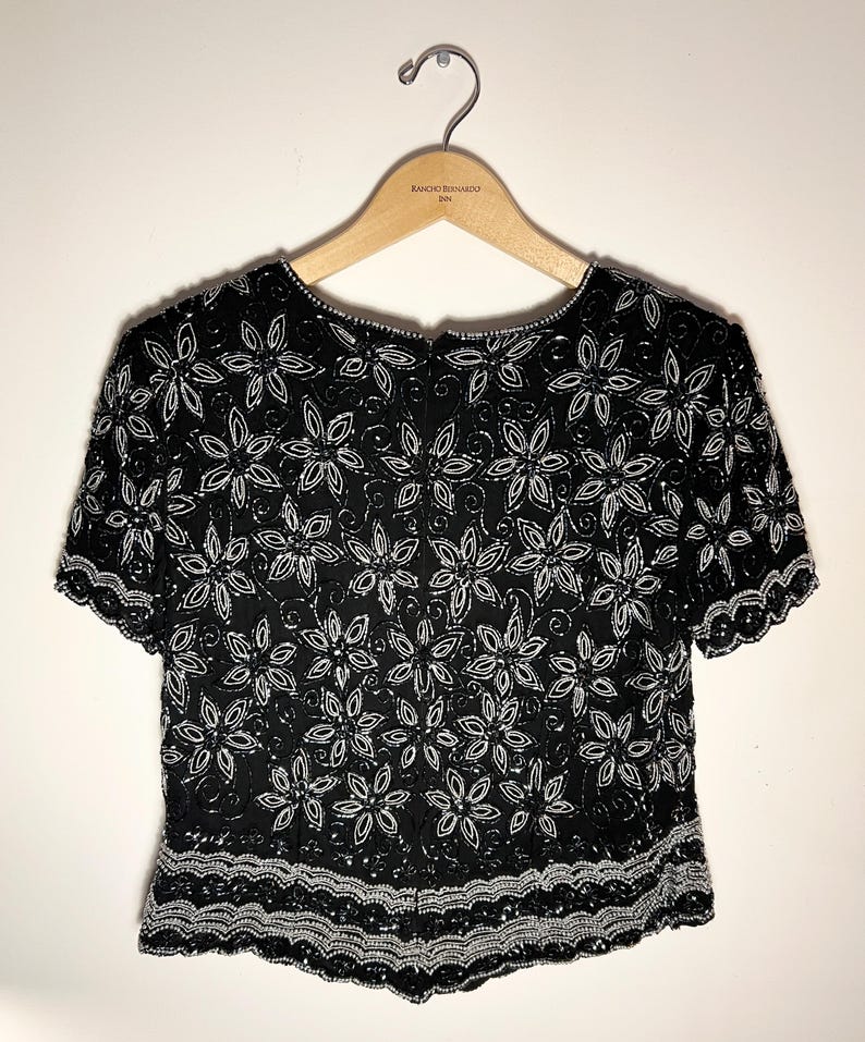 May include: Black beaded top with white floral design. The short-sleeved top is embellished with sequins and beads, featuring a scalloped hemline. The top is hanging on a wooden hanger.