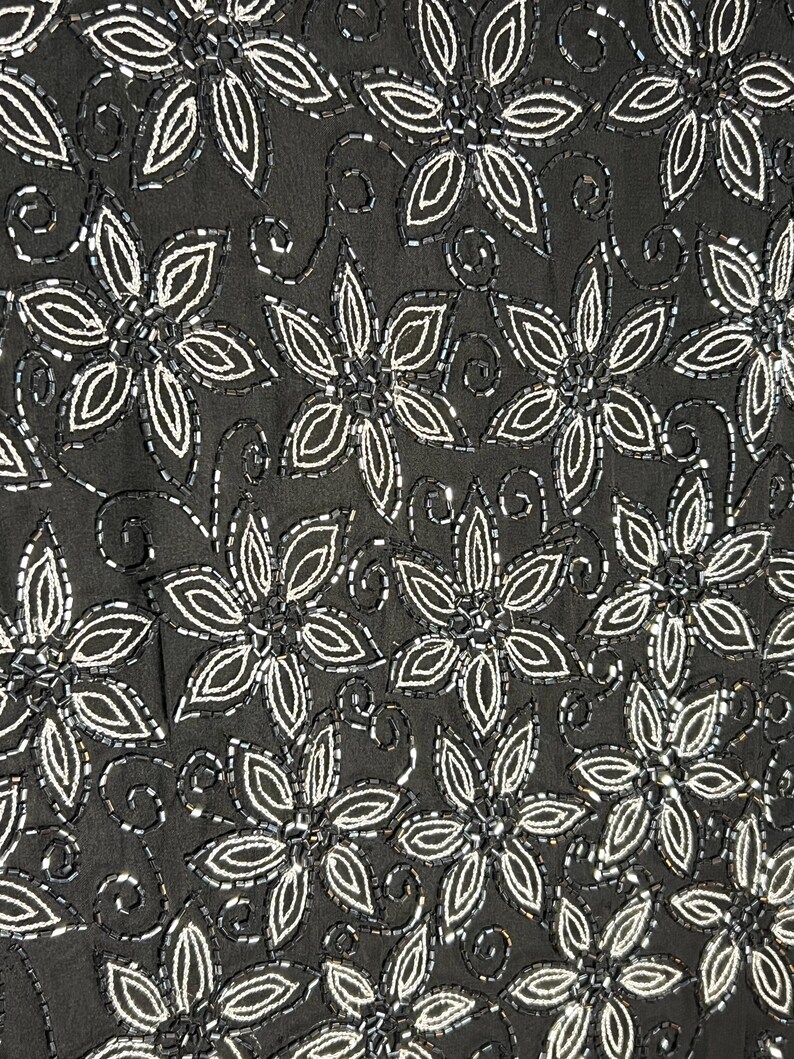May include: Black fabric featuring a repeating floral design. Each flower is outlined and filled with white beads. The background is a solid black colour.