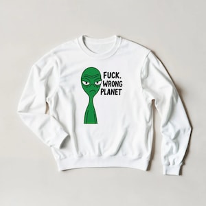 Puede incluir: Sudadera blanca con un diseño de alienígena verde y el texto "FUCK. WRONG PLANET". El alienígena tiene una expresión de enfado. La sudadera es de estilo casual y cómodo.