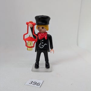Könnte beinhalten: Eine Playmobil-Figur eines Schornsteinfegers, mit schwarzem Hut, Jacke und Hose sowie einem roten Schal. Die Figur hält eine rote Laterne mit goldenem Licht. Die Figur steht auf einer transparenten Kunststoffbasis. Ein kleines weißes Etikett mit der Nummer 396 befindet sich im Vordergrund.