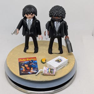May include: Two Playmobil figures in black suits, white shirts, and ties, each holding a silver gun. A miniature Pulp Fiction movie poster, a burger, and a drink are on a circular base. The number 601 is printed on a white label.