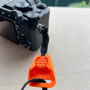 Puede incluir: Primer plano de una cámara negra con un conector de correa naranja. El conector es de color naranja brillante y tiene una superficie texturizada. La correa de la cámara es negra y está sujeta a la cámara.
