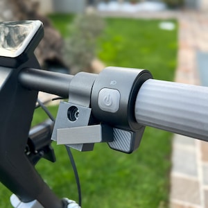 Könnte beinhalten: Nahaufnahme eines Elektroroller-Lenkers. Der Lenker ist schwarz mit einem grauen Griff. Ein graues Bedienfeld zeigt einen Ein-/Ausschalter und ein Scheinwerfersymbol. Eine graue Halterung ist am Lenker befestigt.