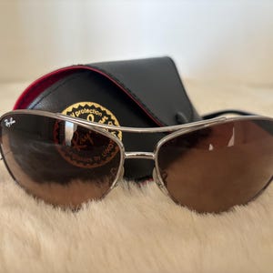 May include: Brown gradient sunglasses with a silver frame and a black case. The sunglasses have a curved design and are resting on a white, fluffy surface. The case is black with a red interior.