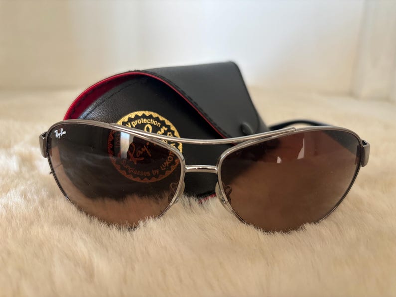 May include: Brown gradient sunglasses with a silver frame and a black case. The sunglasses have a curved design and are resting on a white, fluffy surface. The case is black with a red interior.
