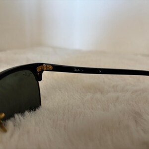 May include: Black sunglasses with gold-coloured accents. The lenses are dark, and the frame is black. The Ray-Ban logo is visible on the arm. The sunglasses are set against a soft, white background.