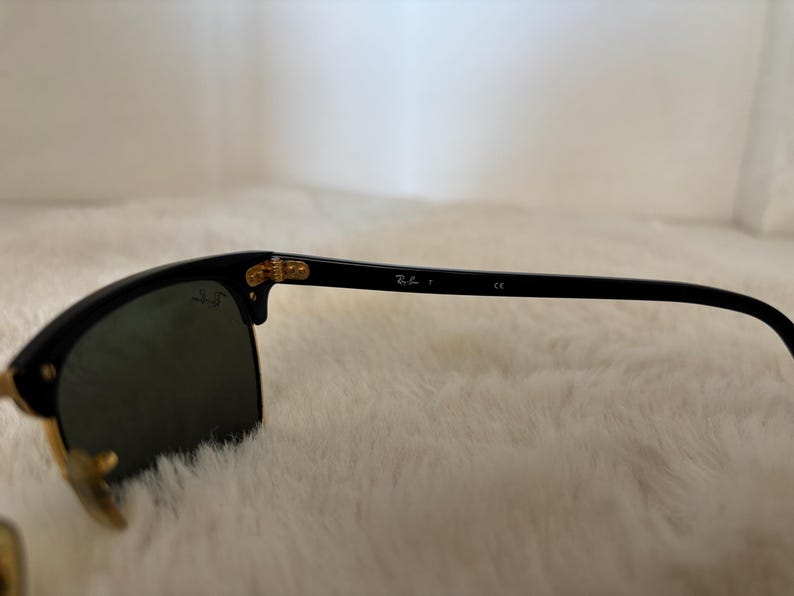 May include: Black sunglasses with gold-coloured accents. The lenses are dark, and the frame is black. The Ray-Ban logo is visible on the arm. The sunglasses are set against a soft, white background.