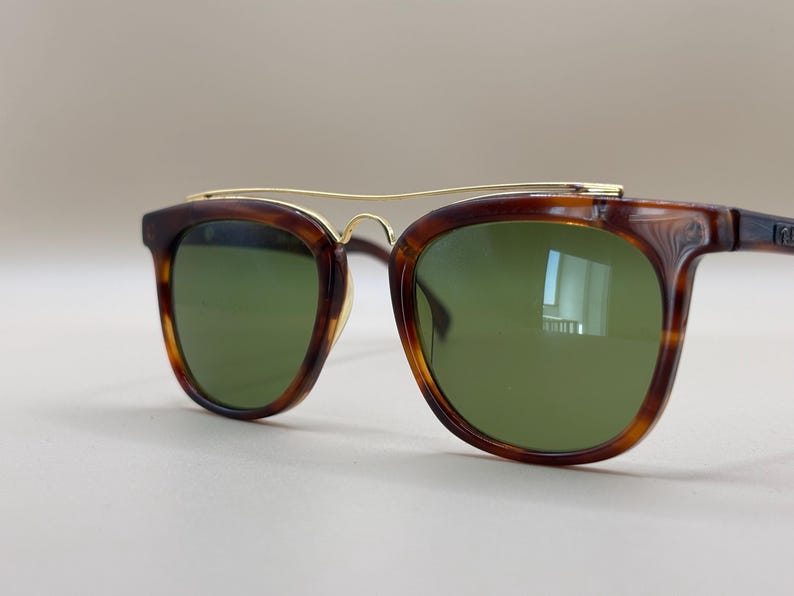 May include: Brown tortoiseshell sunglasses with green lenses. The sunglasses have a gold-colored metal bridge and arms. The frames are a classic shape, suitable for various face shapes. The lenses are a solid green color.