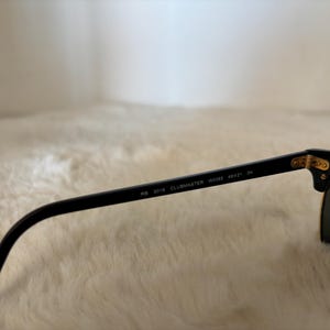 May include: Black sunglasses with the text "RB 3016 CLUBMASTER W0366 49[]21 3N" on the arm. The sunglasses have a gold-coloured accent at the hinge and green lenses. The sunglasses are resting on a white, fluffy surface.