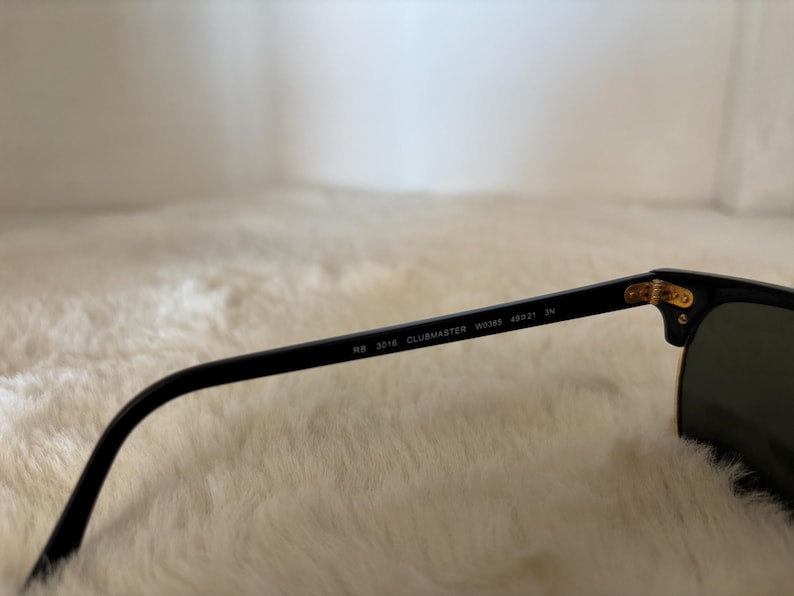 May include: Black sunglasses with the text "RB 3016 CLUBMASTER W0366 49[]21 3N" on the arm. The sunglasses have a gold-coloured accent at the hinge and green lenses. The sunglasses are resting on a white, fluffy surface.