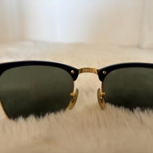 May include: Black and gold sunglasses with green lenses. The sunglasses feature a classic design with a black upper frame and gold accents. The lenses are a dark green colour, and the frame has a gold bridge and hinges.