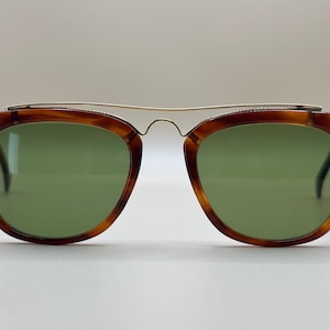 May include: Vintage sunglasses with a tortoiseshell frame and green lenses. The glasses have a gold-colored metal bridge that connects the two lenses. The frames are a warm brown color with darker marbling.