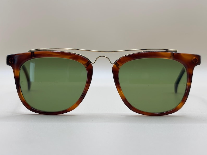 May include: Vintage sunglasses with a tortoiseshell frame and green lenses. The glasses have a gold-colored metal bridge that connects the two lenses. The frames are a warm brown color with darker marbling.