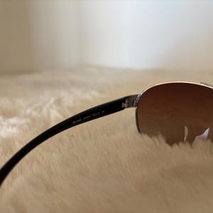 May include: Aviator-style sunglasses with a silver frame and brown gradient lenses. The black temples have the text "RB 3386 004/13 59/13 3N". The sunglasses are resting on a soft, white surface.