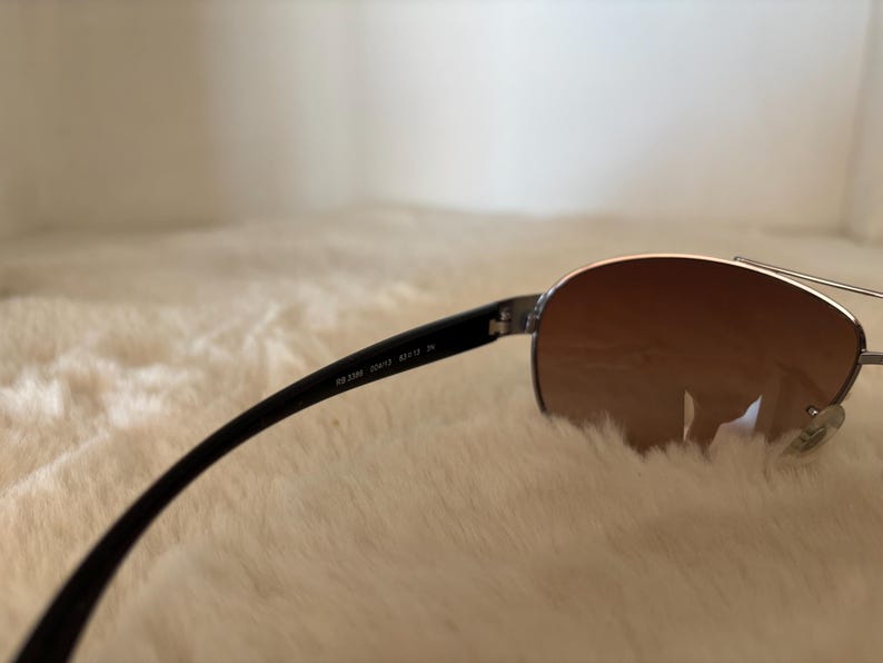 May include: Aviator-style sunglasses with a silver frame and brown gradient lenses. The black temples have the text "RB 3386 004/13 59/13 3N". The sunglasses are resting on a soft, white surface.