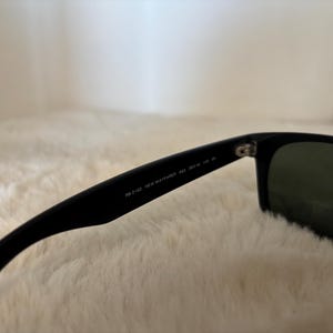 K&ouml;nnte beinhalten: Schwarze Sonnenbrille mit dunkelgr&uuml;nen Gl&auml;sern. Auf dem B&uuml;gel steht "RB 2132 NEW WAYFARER 52 18 145". Die Sonnenbrille liegt vor einem weichen, wei&szlig;en Hintergrund.