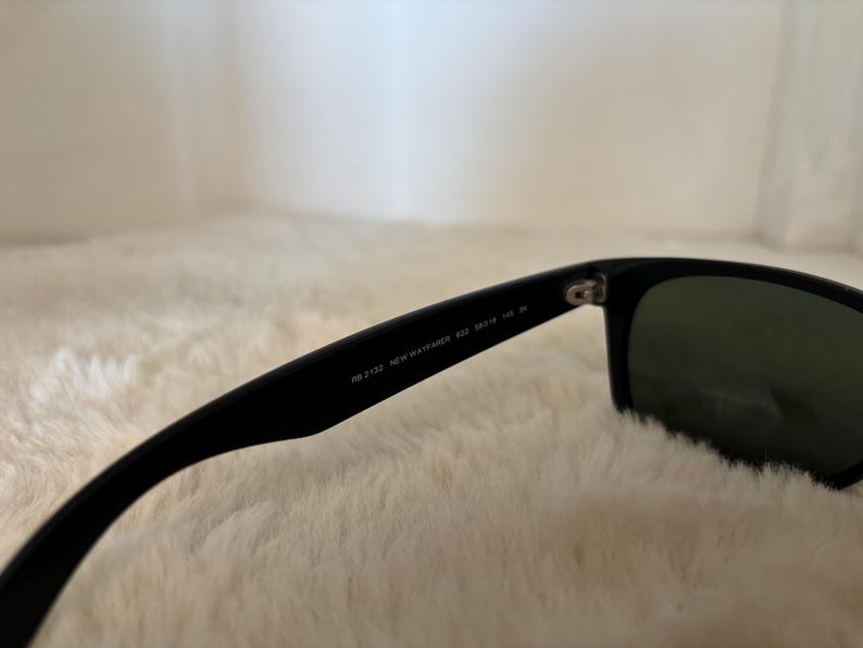 K&ouml;nnte beinhalten: Schwarze Sonnenbrille mit dunkelgr&uuml;nen Gl&auml;sern. Auf dem B&uuml;gel steht "RB 2132 NEW WAYFARER 52 18 145". Die Sonnenbrille liegt vor einem weichen, wei&szlig;en Hintergrund.