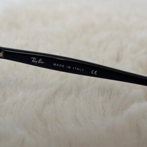 May include: Close-up of a black Ray-Ban sunglass arm with the brand name and "MADE IN ITALY" printed in white. The arm also has a "CE" marking. The background is a soft, white, textured surface.