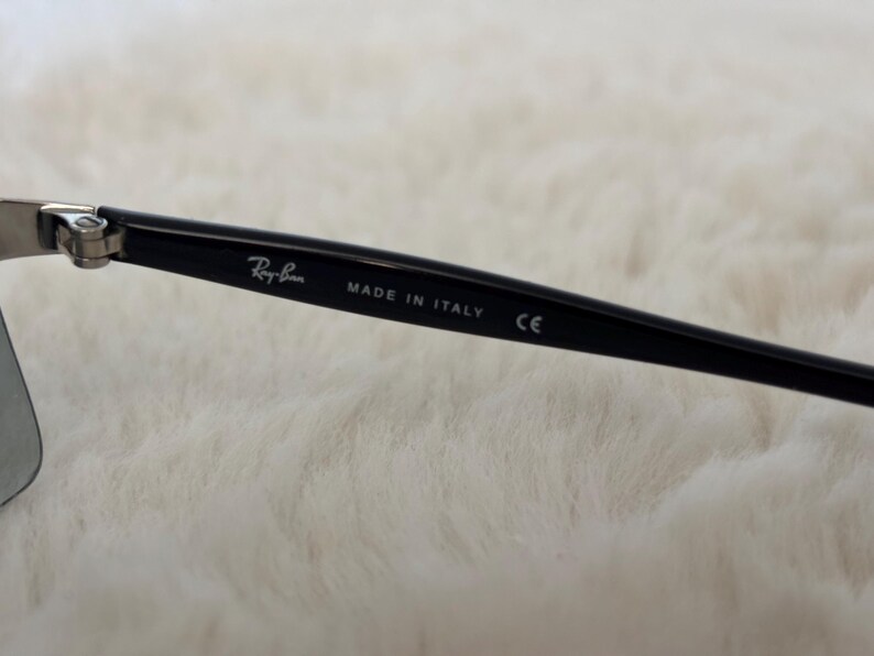 May include: Close-up of a black Ray-Ban sunglass arm with the brand name and "MADE IN ITALY" printed in white. The arm also has a "CE" marking. The background is a soft, white, textured surface.